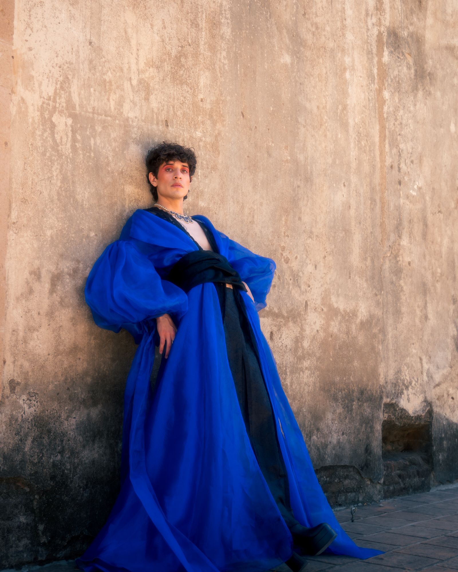 A male model in a blue gown leaning against a textured wall.